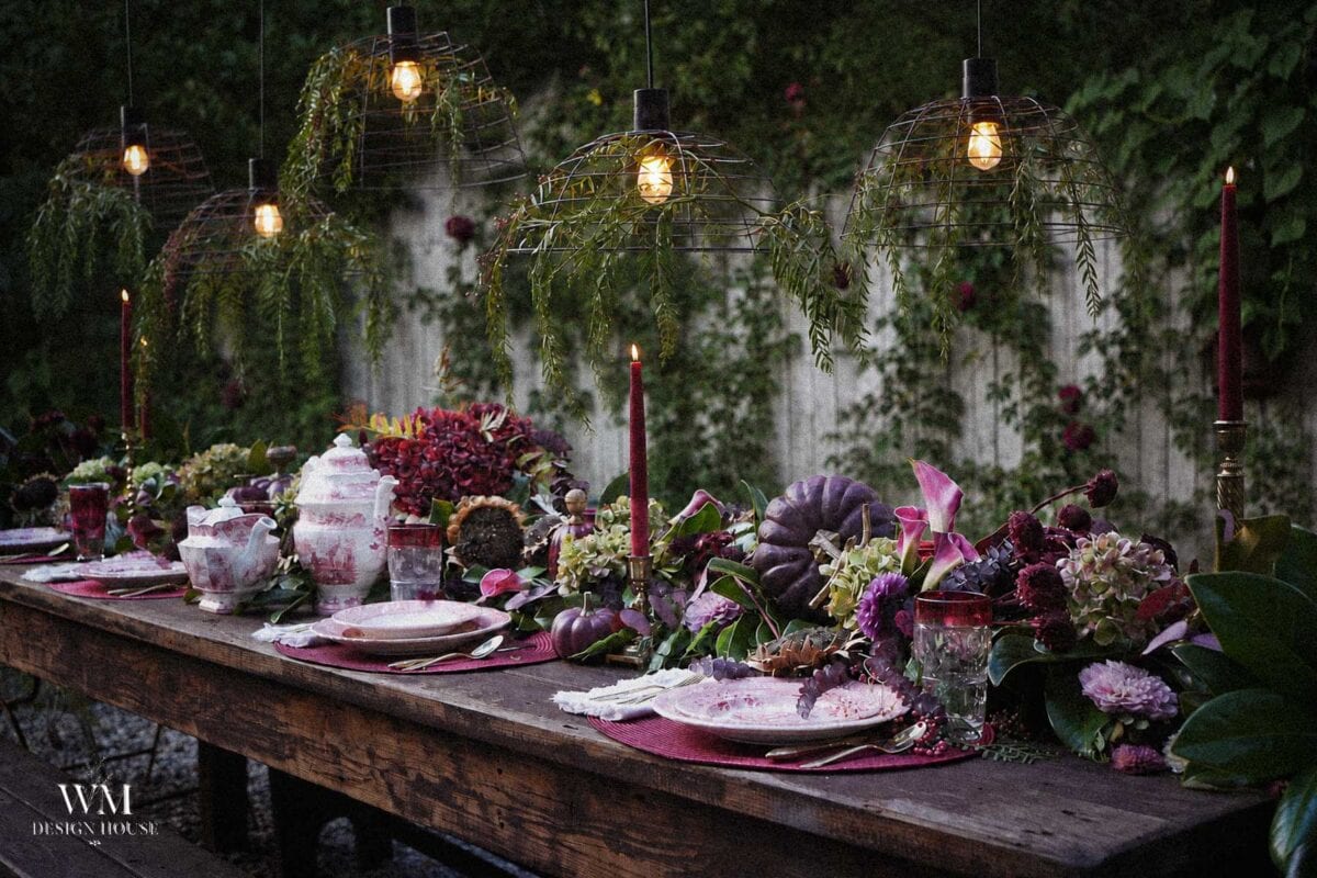 A rustic outdoor table set with plates, flowers, candles, and pumpkins under hanging lights with greenery, against a vine-covered wall.
