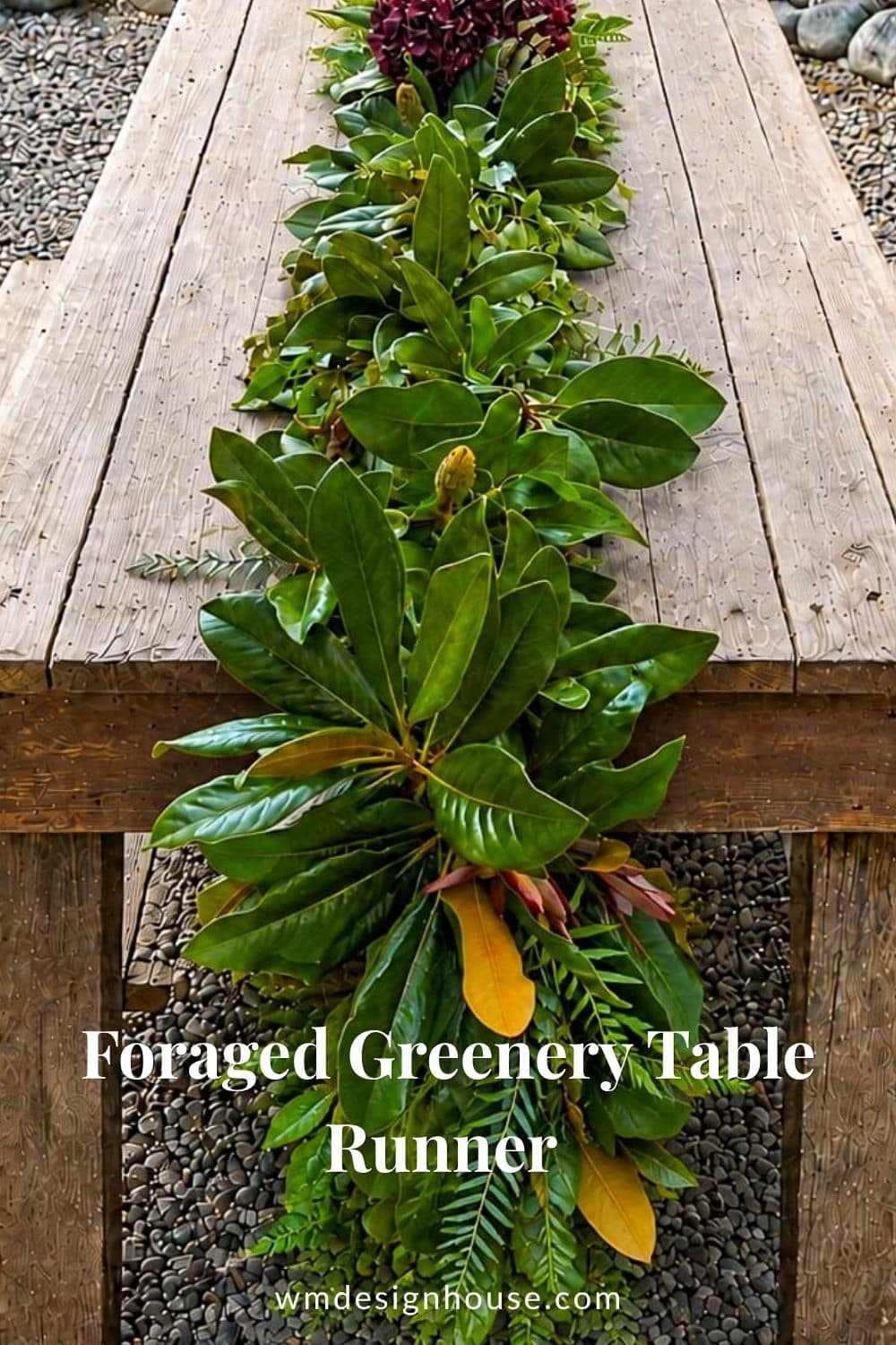 A rustic wooden table with a lush greenery table runner made of assorted leaves and branches, placed outdoors on a stone surface. Text reads: “Foraged Greenery Table Runner” and “wmdesignhouse.com”.