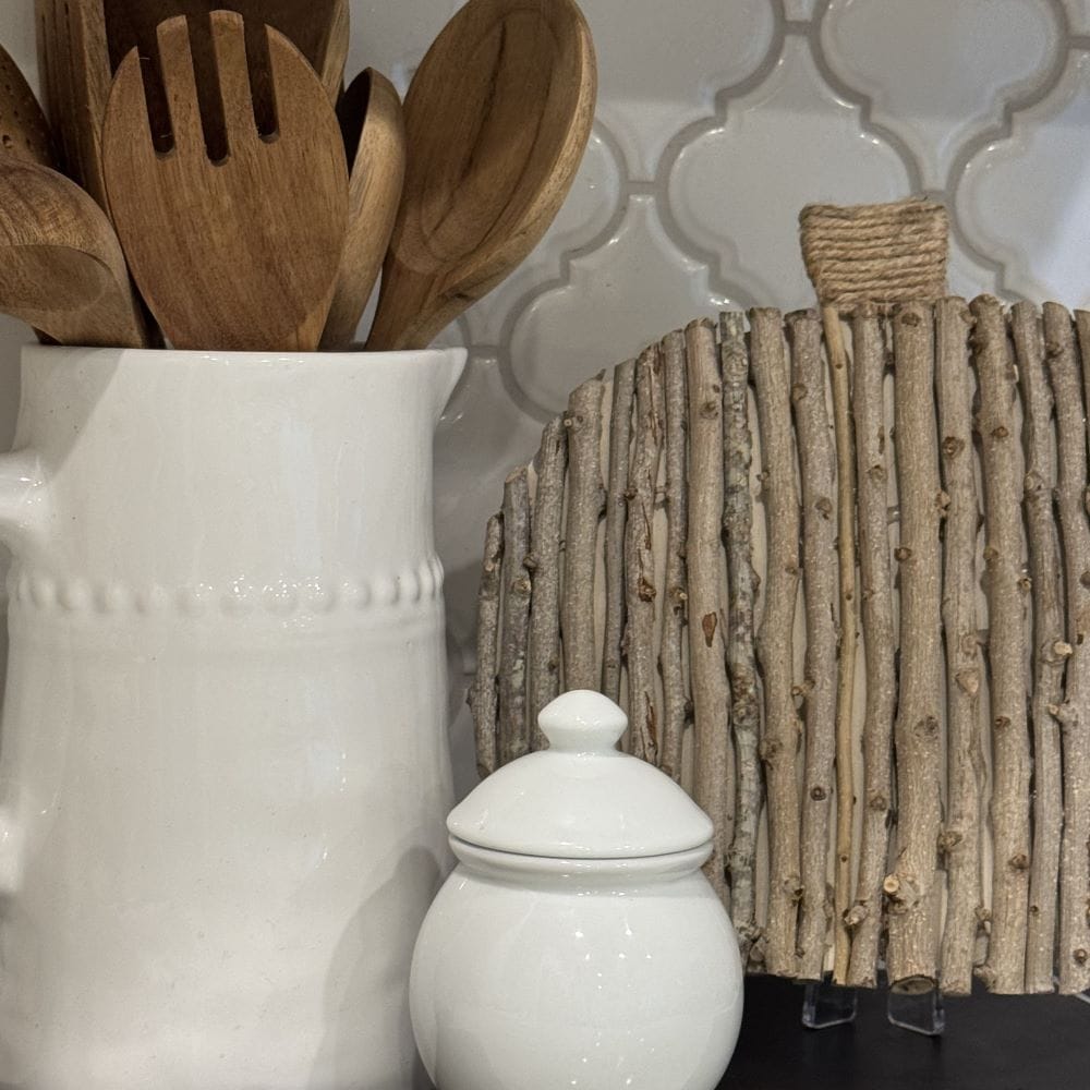 White ceramic pitcher holding wooden utensils, a small white jar with a lid, and a decorative cutting board made of vertical sticks, set against a white tiled backsplash.