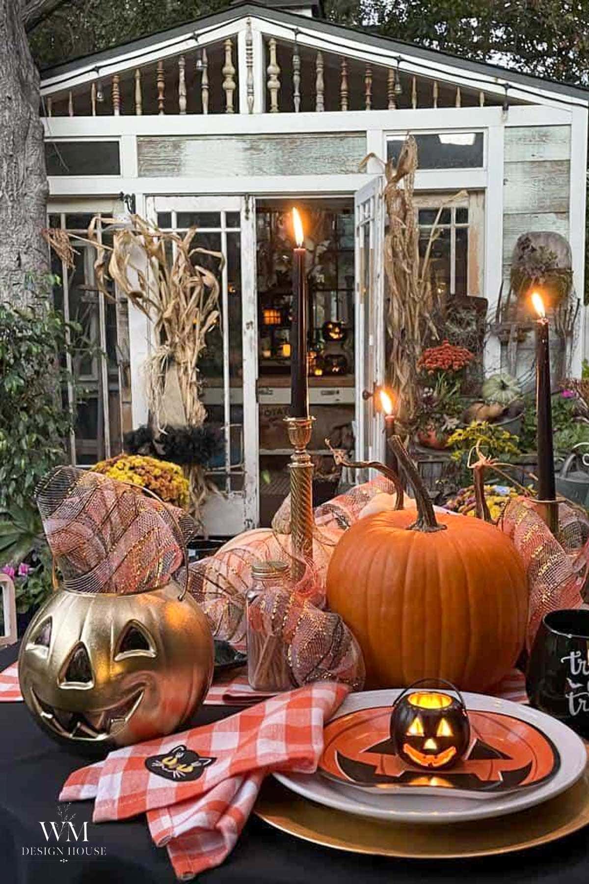 A Halloween-themed outdoor table with pumpkins, candles, mesh ribbon, and festive plates, set in front of a glass garden house decorated with corn stalks and autumn plants.