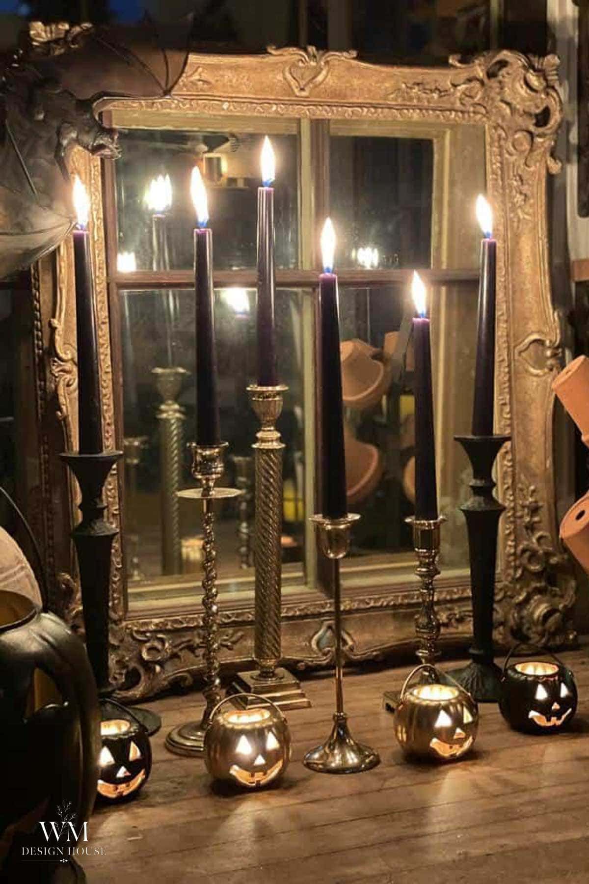 Five tall black candles in ornate holders stand before a large antique mirror. Three small metallic pumpkins with carved faces sit at the base, their reflections creating a Halloween-themed, moody atmosphere.