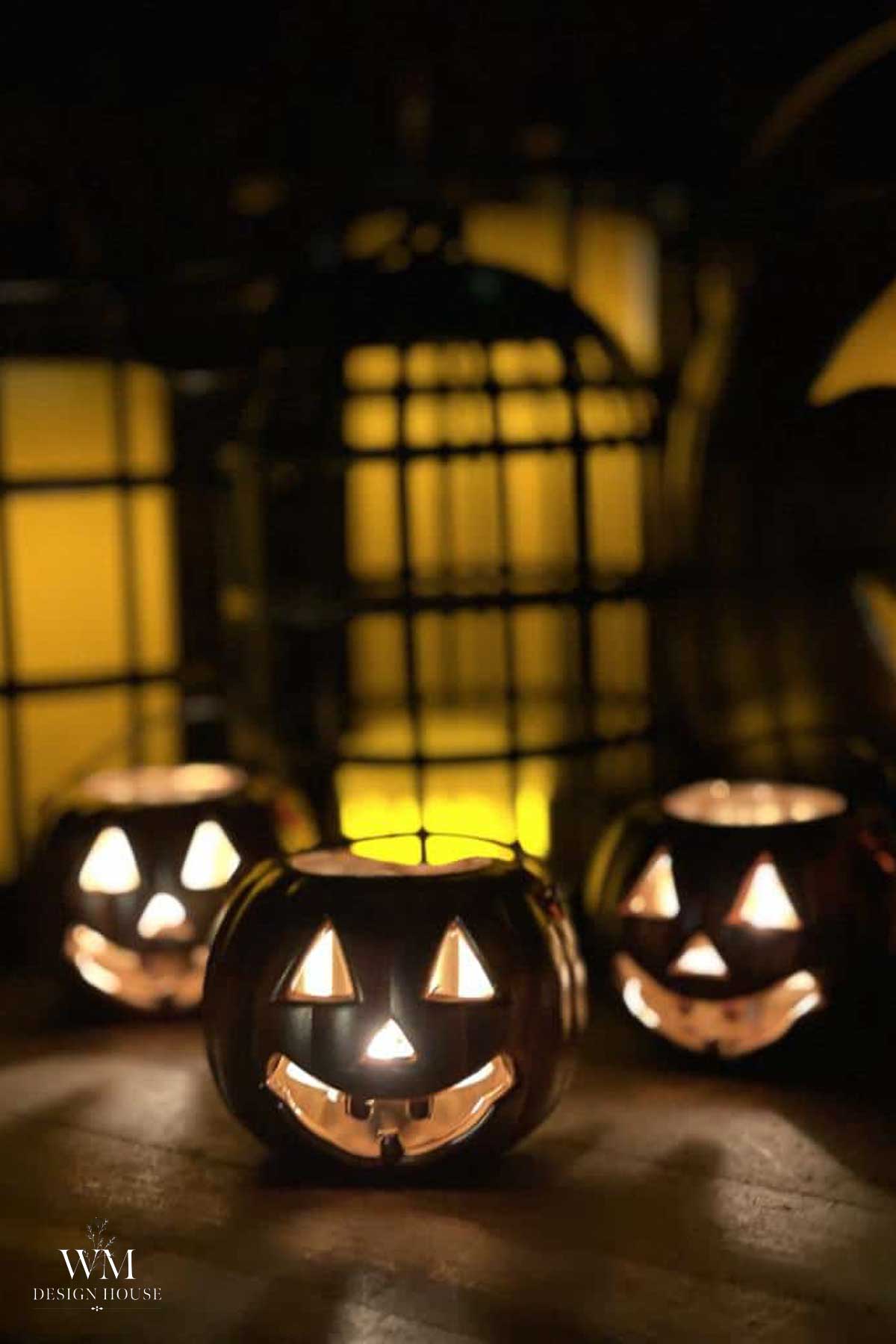 Three glowing jack-o’-lanterns with smiling faces sit on a dark surface, illuminated by candles inside. A yellowish light in the background adds a warm, spooky Halloween atmosphere.