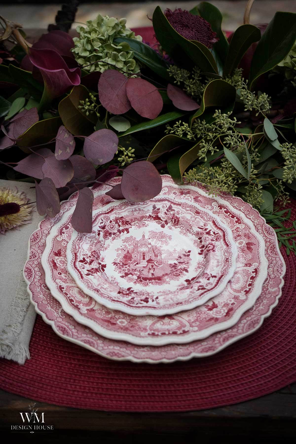 Three vintage-style red and white plates are stacked on a red placemat, next to a beige napkin and surrounded by assorted greenery and purple-toned flowers.
