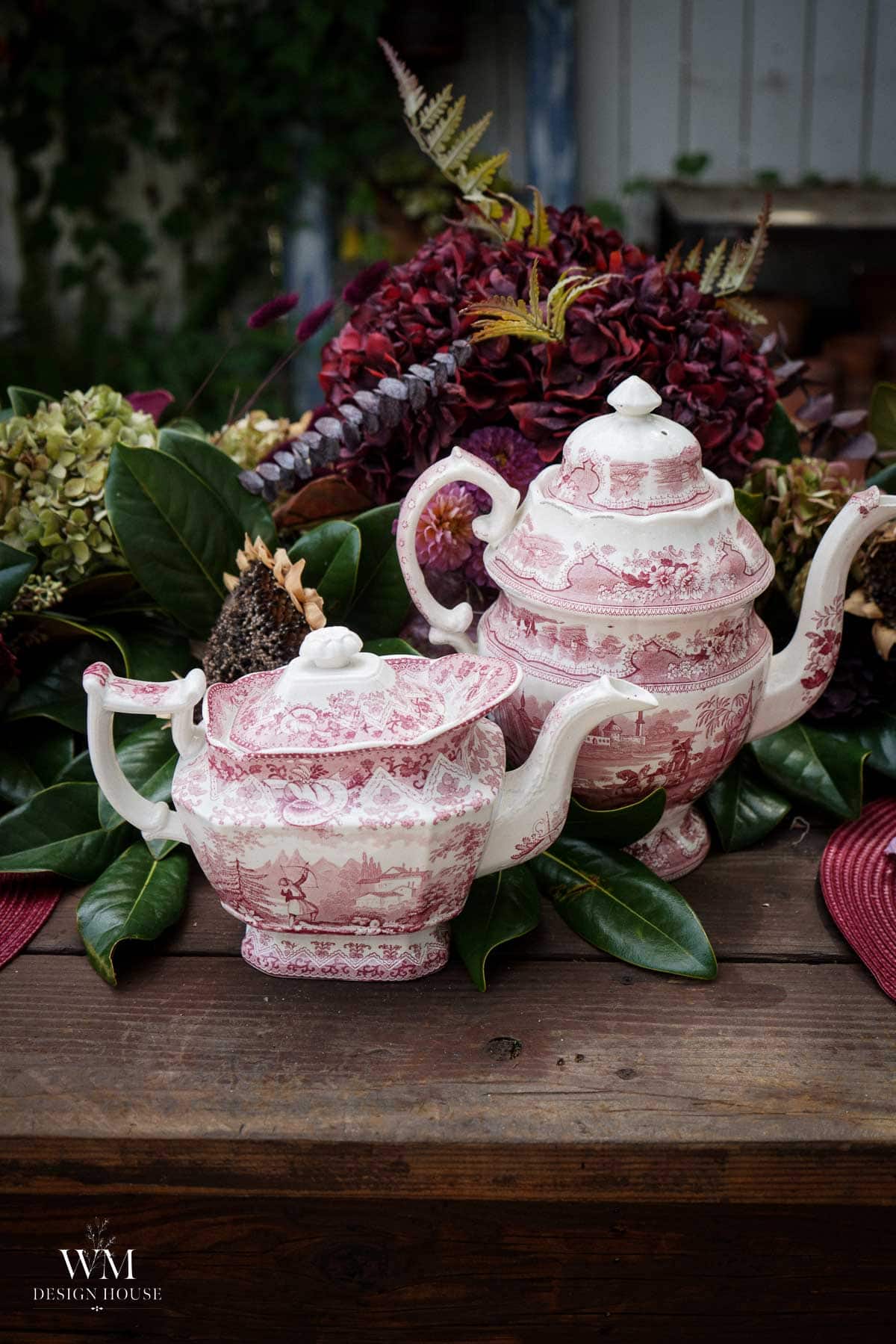 Two ornate, pink-and-white teapots with floral patterns sit on a wooden table, surrounded by green leaves and red, purple, and brown flowers as decoration.