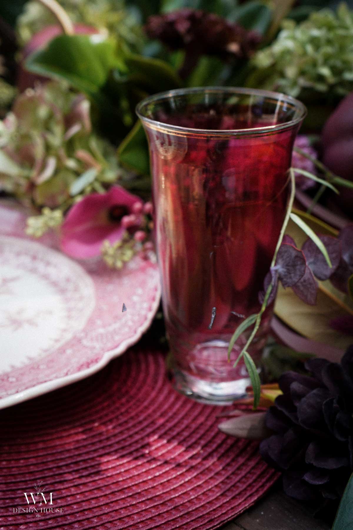 A glass of red drink sits on a pink woven placemat beside a floral-patterned plate and an arrangement of fresh flowers in pink and green hues.