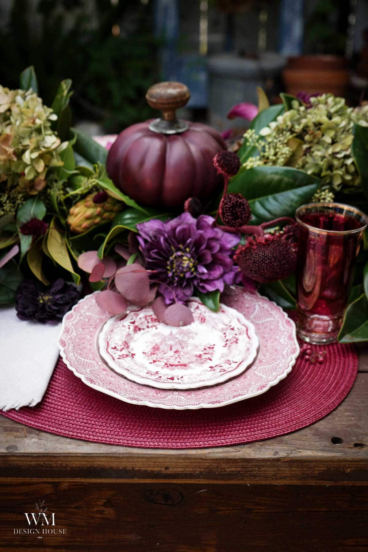 Elegant table setting with vintage pink plates on a pink placemat, surrounded by purple flowers, leafy greenery, a decorative purple pumpkin, and a glass filled with a red drink.