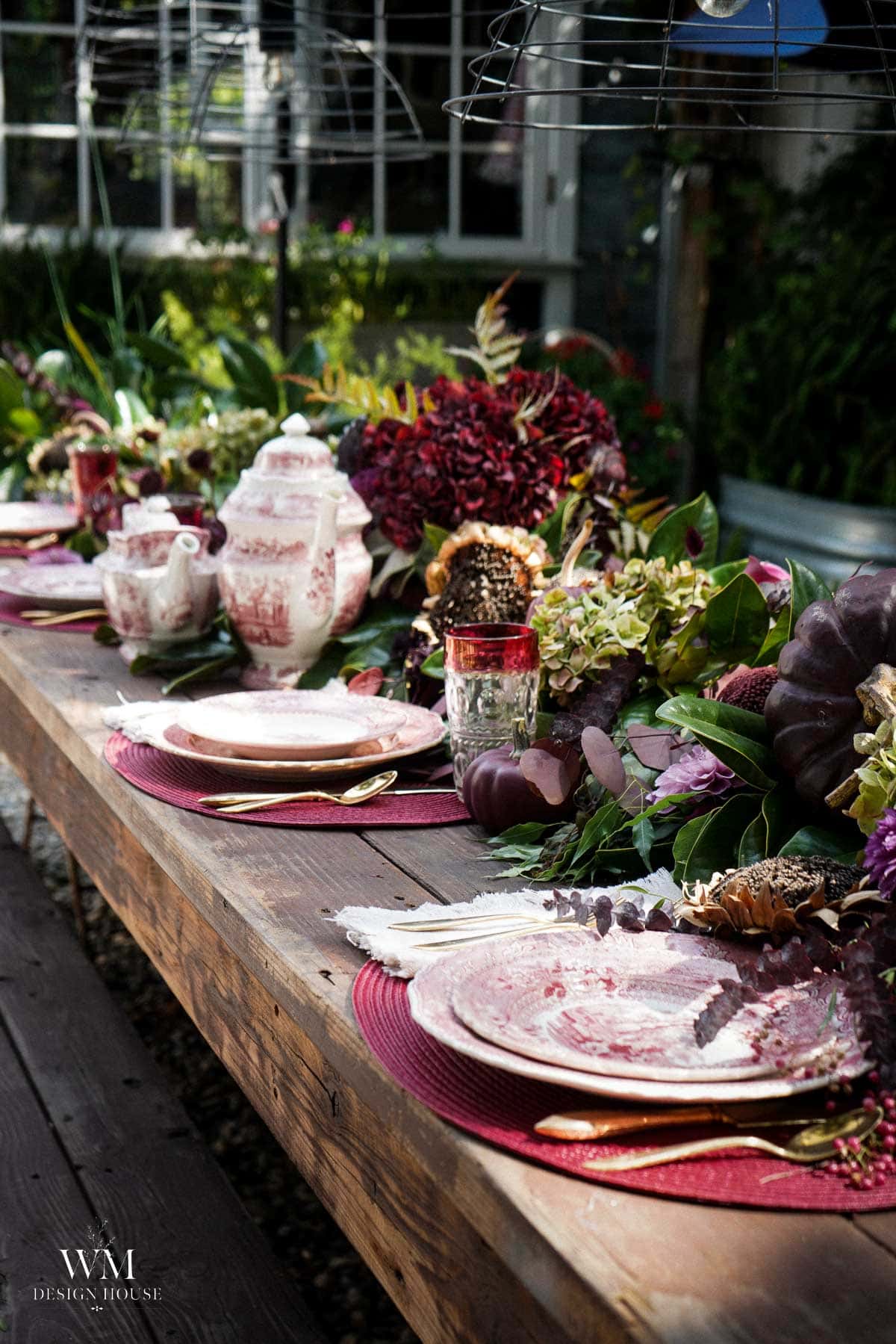 An outdoor wooden table is set with floral china, gold cutlery, dark red and pink flowers, and greenery for a garden party. Sunlight filters through, creating a cozy, elegant atmosphere.