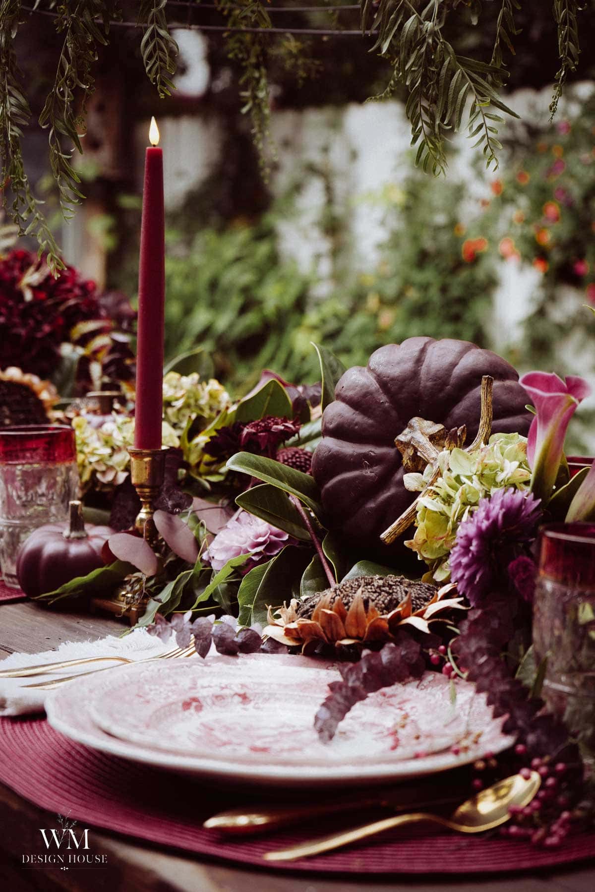 A rustic outdoor table is set with burgundy candles, dark purple pumpkins, assorted flowers, greenery, and elegant plates, creating an autumnal, festive atmosphere.