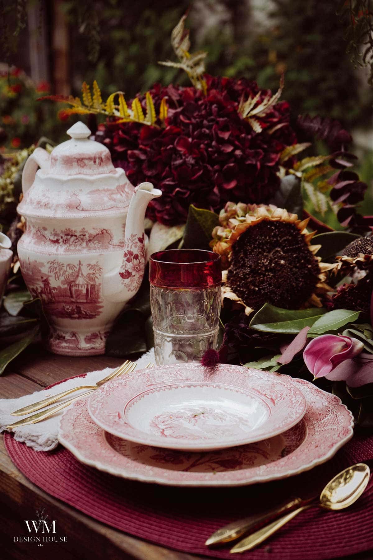 A vintage tea set with pink patterns, gold cutlery, and a glass sit on a wooden table with burgundy flowers and sunflowers, creating an elegant, rustic outdoor setting.