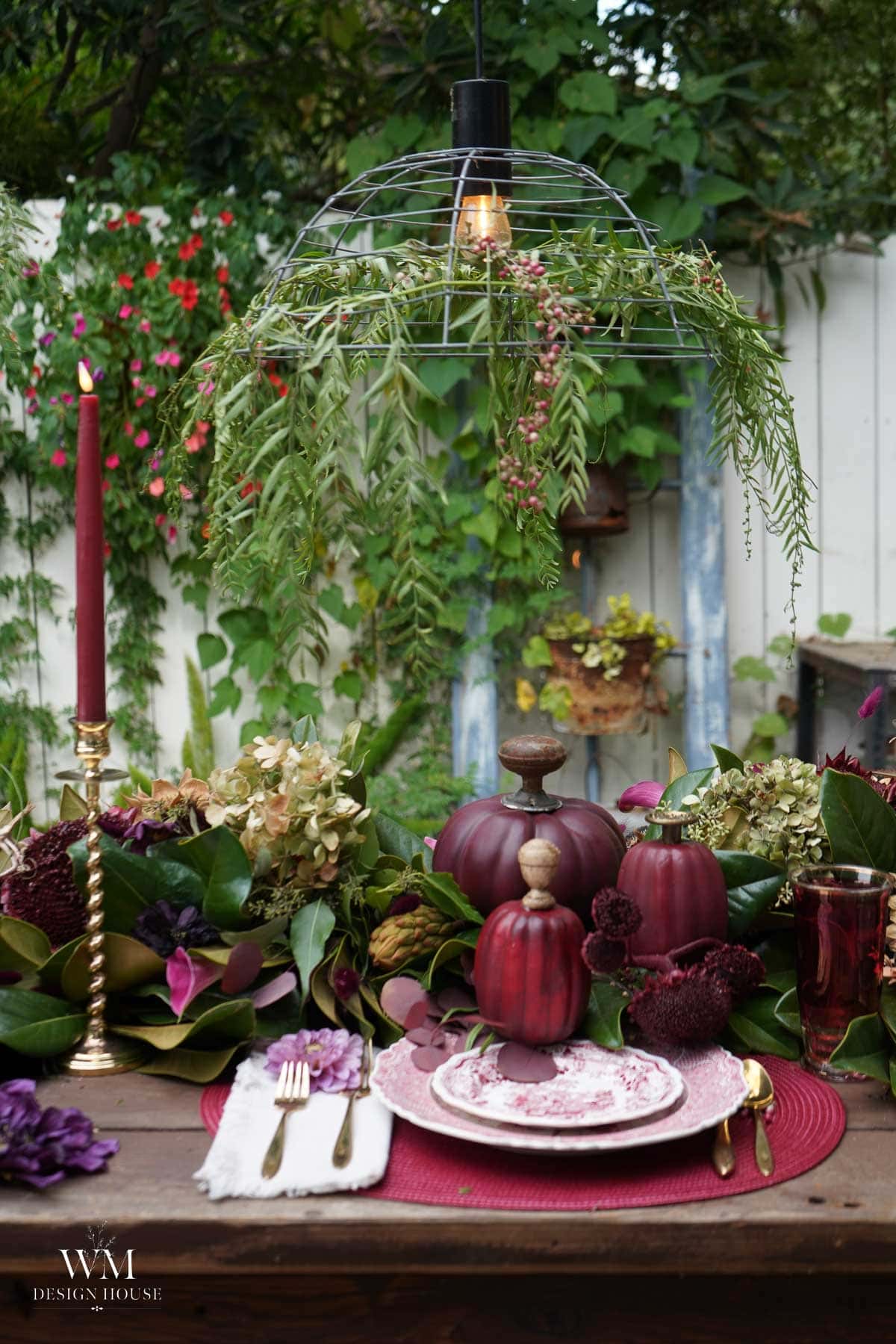 A rustic outdoor table is set with burgundy pumpkins, floral arrangements, red candles, and vintage plates. Greenery hangs from a wire light fixture above, creating an elegant, autumn-inspired dining scene.