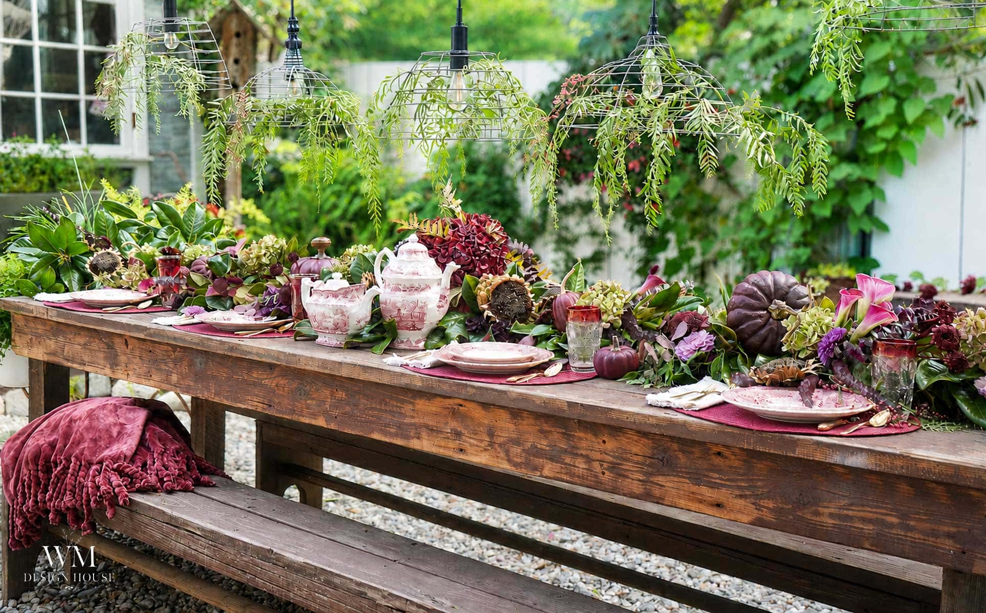 A rustic wooden table set outdoors with pink dishes, decorative gourds, lush greenery, and hanging plants, creating an elegant and vibrant garden dining scene.