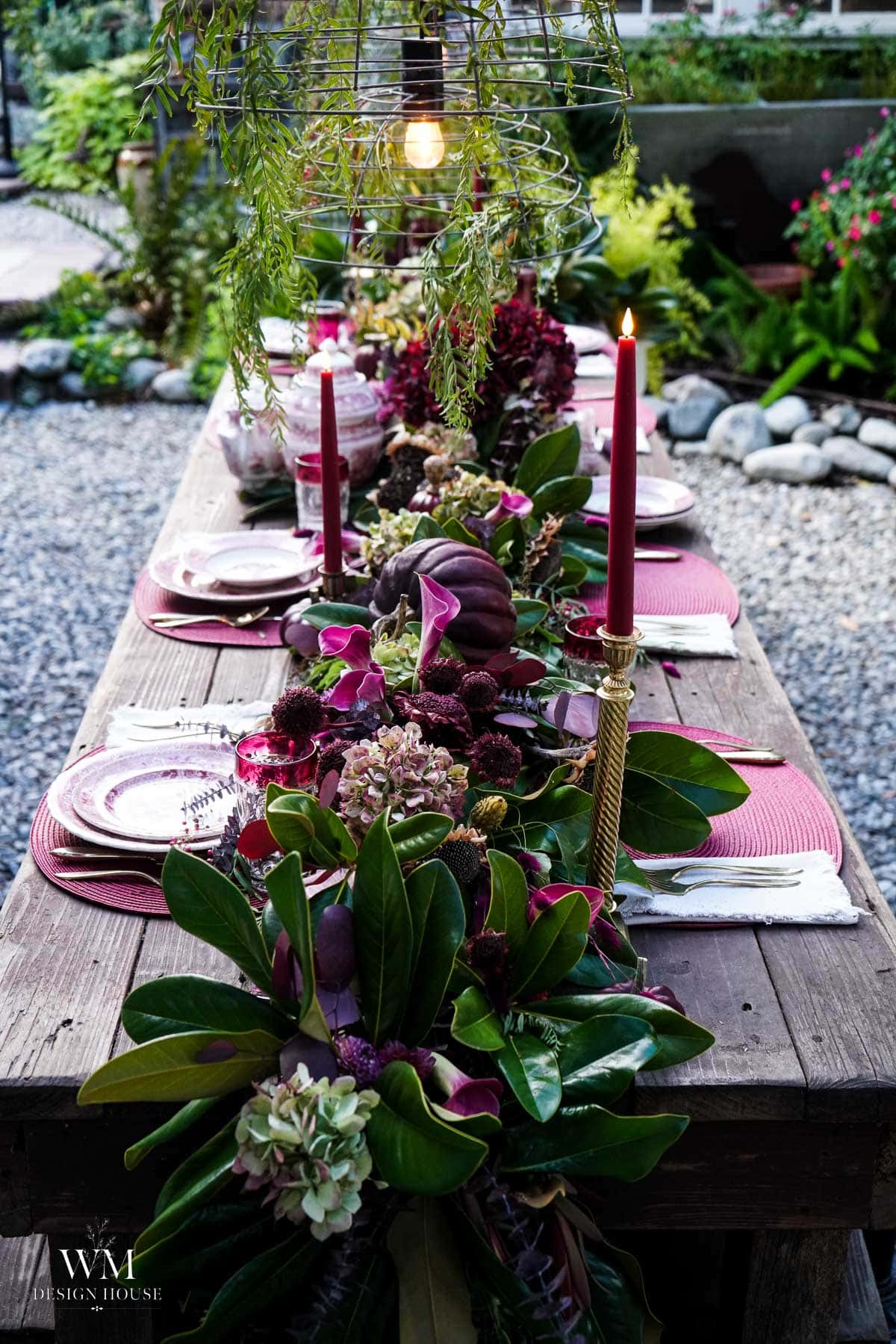 A rustic wooden table is set outdoors with pink plates, gold utensils, red candles, and a lush floral and greenery centerpiece, creating an elegant, inviting garden dining scene.