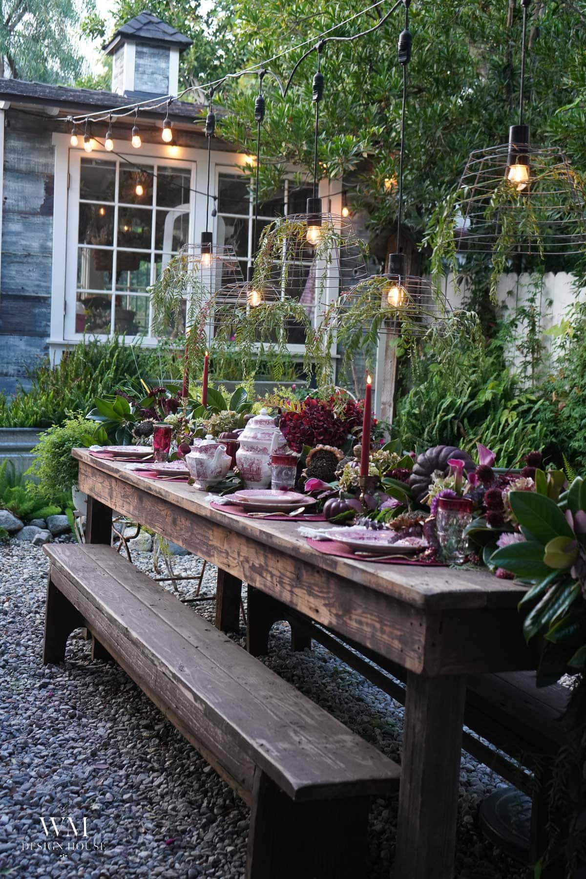 A rustic outdoor dining table set for a meal with candles, china, and flowers, surrounded by greenery and string lights hanging above in a garden setting.