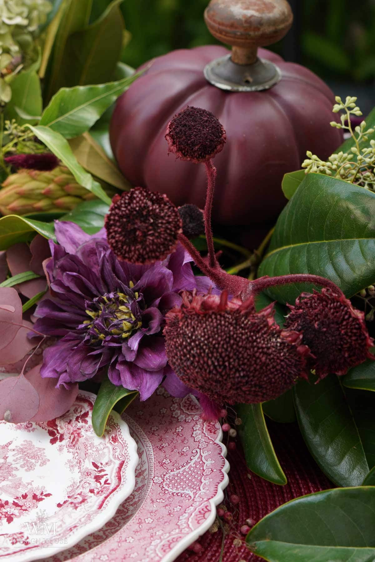 A close-up of a table decorated with a purple pumpkin, dark red flowers, green leaves, and pink patterned plates. The arrangement has a rich, autumnal feel.
