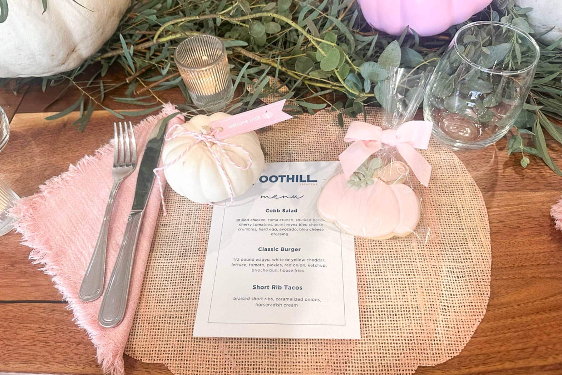 A rustic table setting with a menu, pink napkin, knife and fork, white pumpkin, heart-shaped cookie, glass, and greenery decor. The theme appears casual and elegant with light pastel accents.