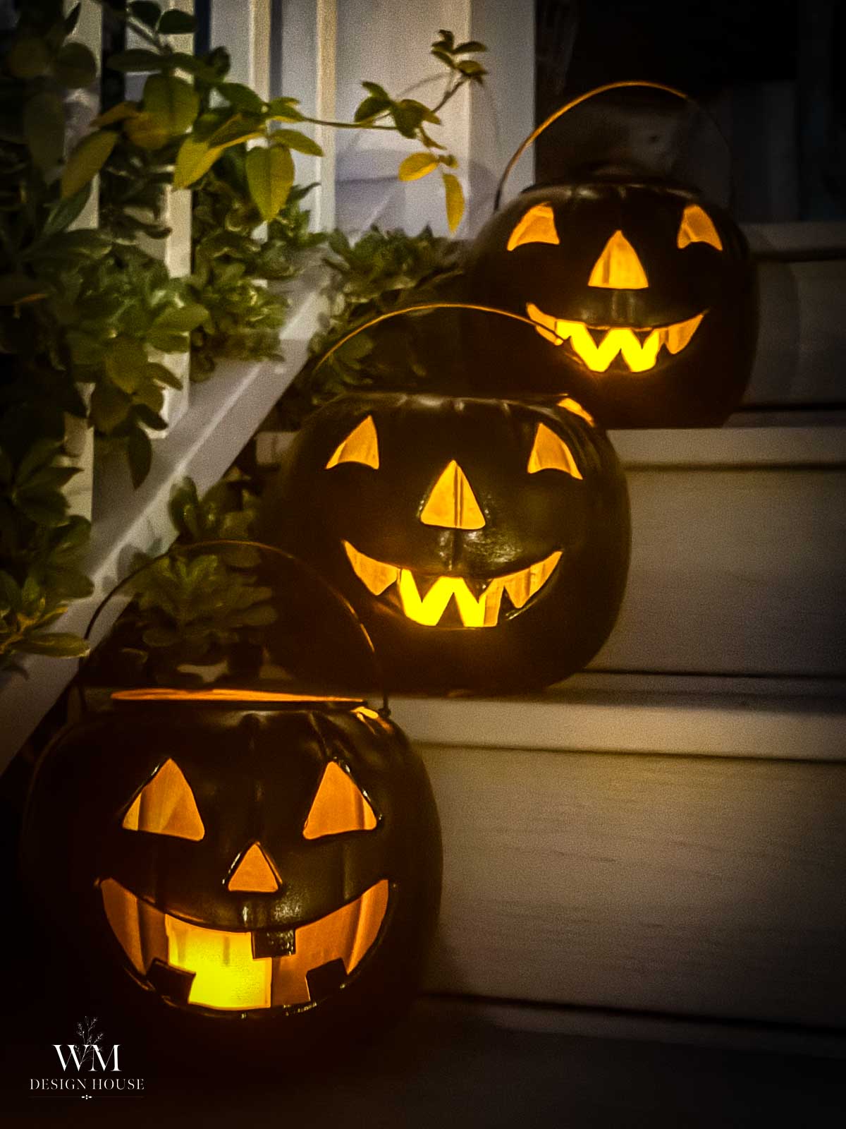 Three glowing jack-o’-lantern buckets with carved faces sit on white stairs at night, surrounded by green leaves, creating a festive Halloween atmosphere and inspiring halloween lanterns DIY ideas.