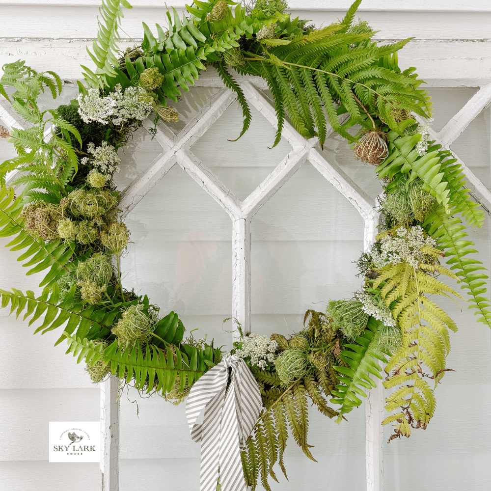 A green fern wreath with white flowers and a striped bow hangs on a glass window with a white frame. The wreath has a natural, fresh look and fills most of the window’s central pane.