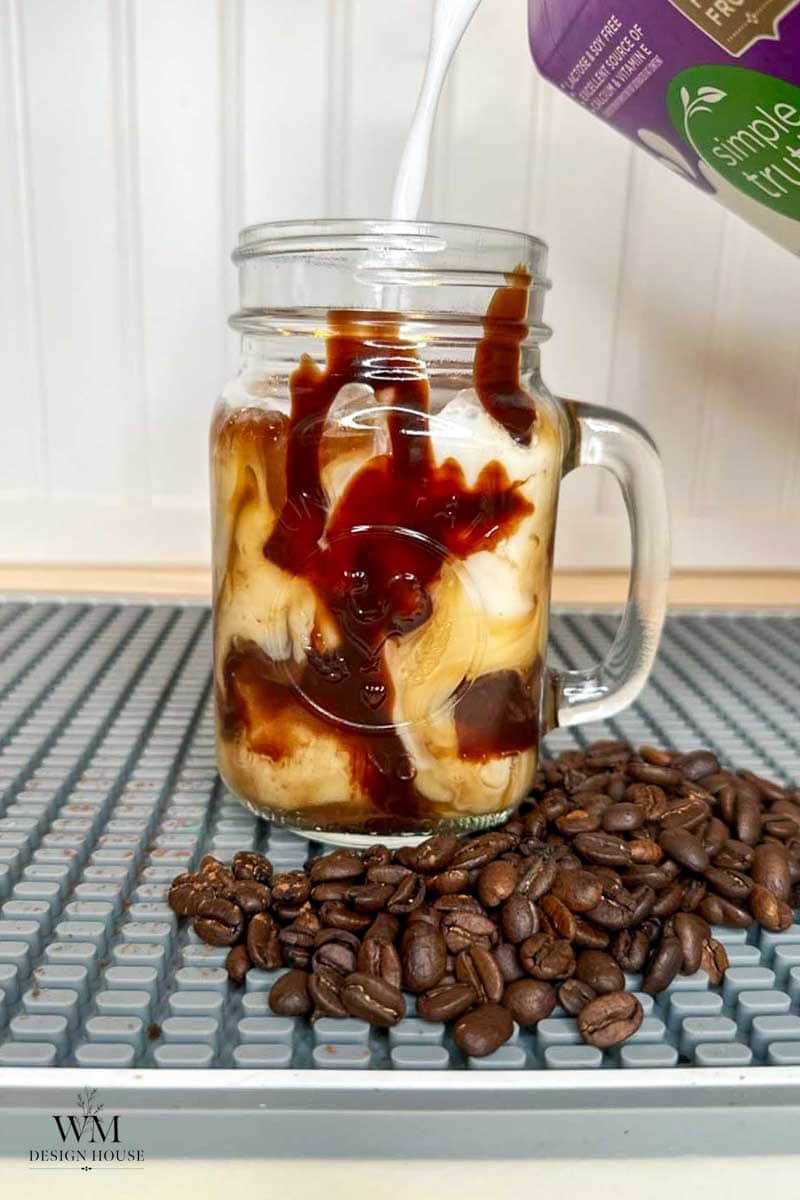 Iced coffee with cream being poured into a mason jar, swirls of chocolate syrup are visible, and coffee beans are scattered in front of the glass.