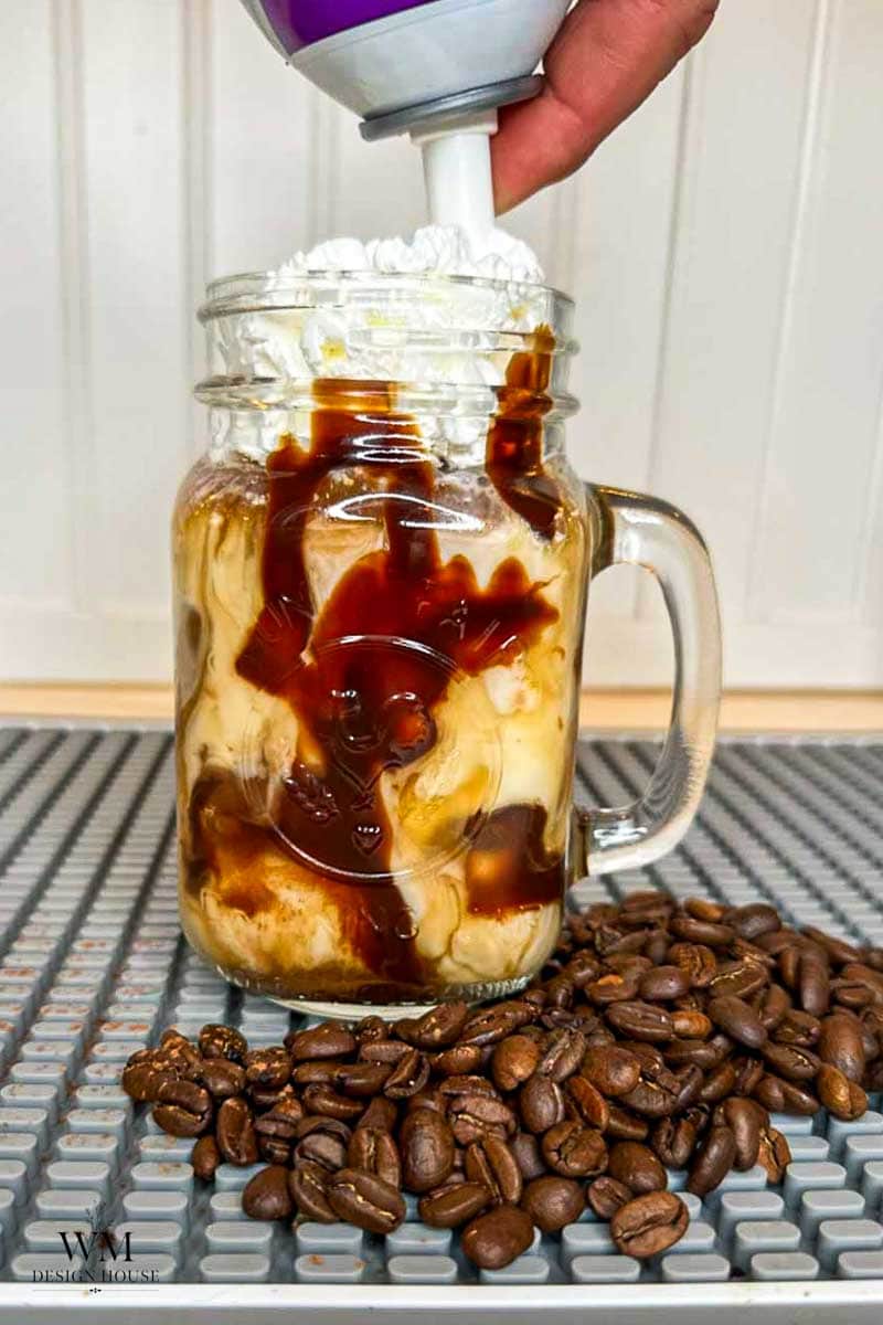 A mason jar of iced coffee with whipped cream and chocolate syrup, next to a pile of coffee beans. A hand is dispensing whipped cream on top.
