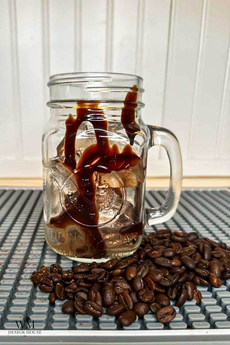 A mason jar filled with ice has coffee being poured into it, with coffee beans scattered on the counter beside the jar.