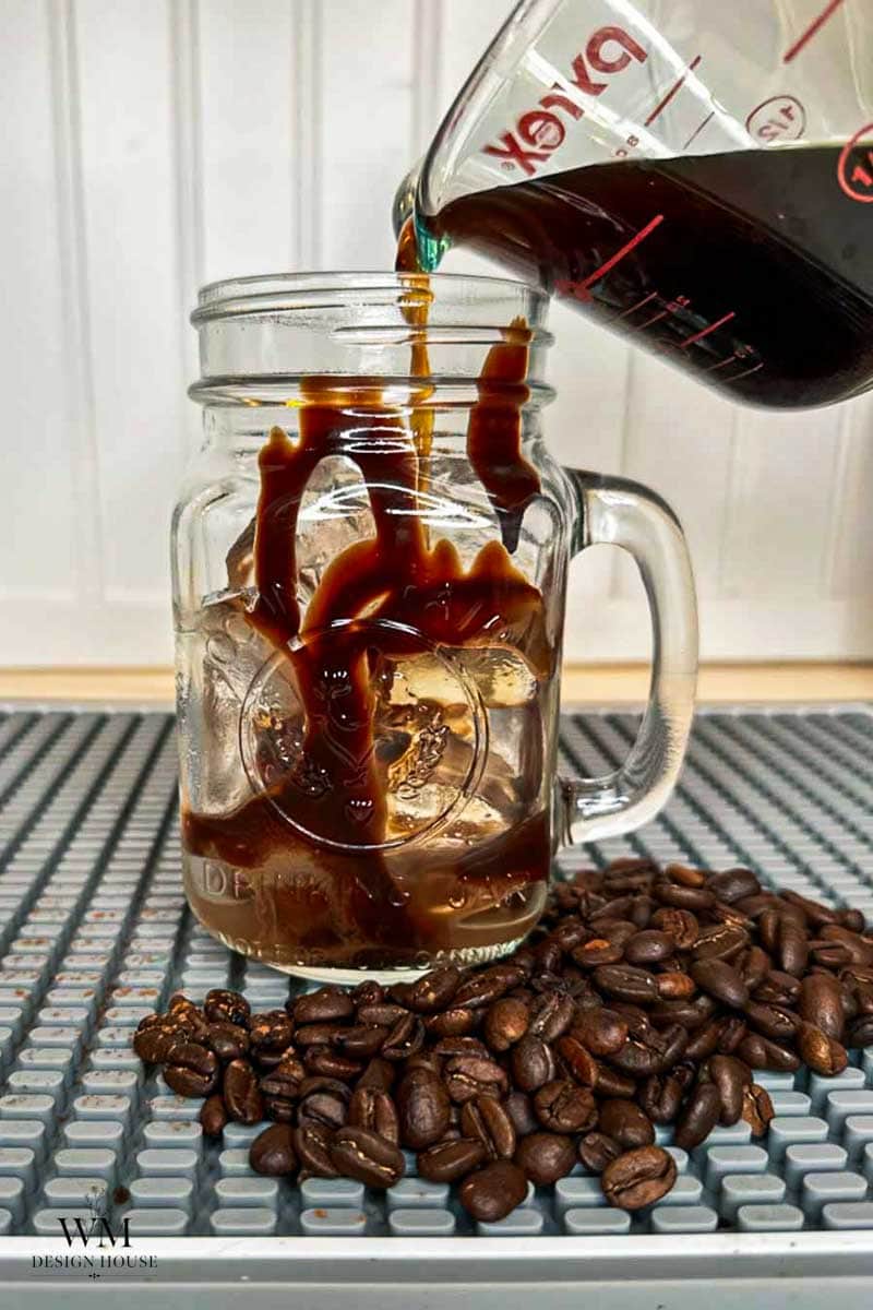 Coffee is being poured from a glass measuring cup into a mason jar filled with ice. Coffee beans are scattered on the surface near the jar.