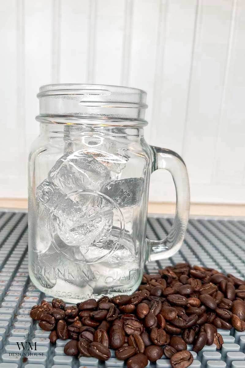 A mason jar filled with large ice cubes sits on a textured surface, with a pile of coffee beans scattered in front of it.