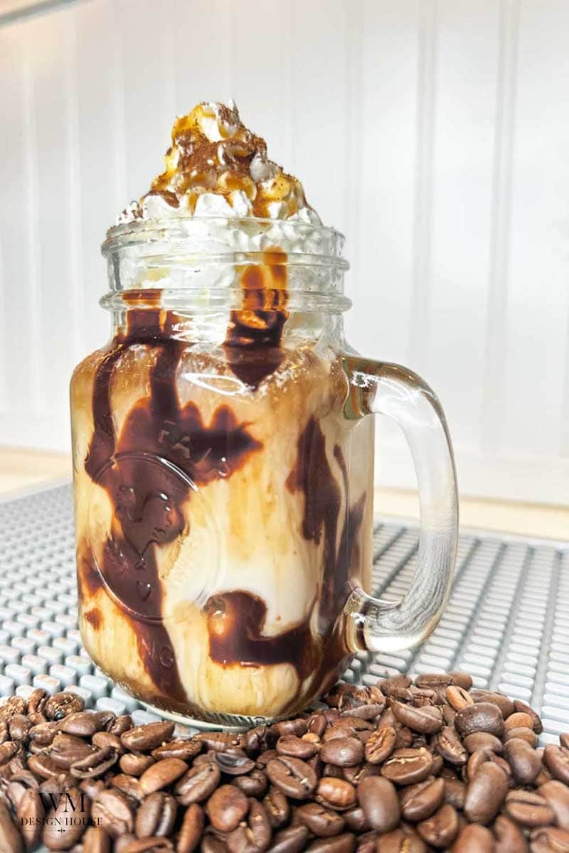 A mason jar filled with iced coffee, whipped cream, and chocolate syrup sits on a metal surface, with coffee beans scattered in front.