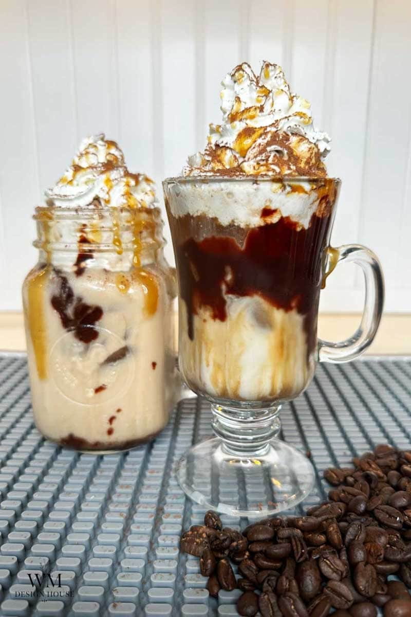 Two glass cups filled with iced coffee drinks topped with whipped cream, chocolate, and caramel drizzle. Coffee beans are scattered in the foreground on a textured surface.
