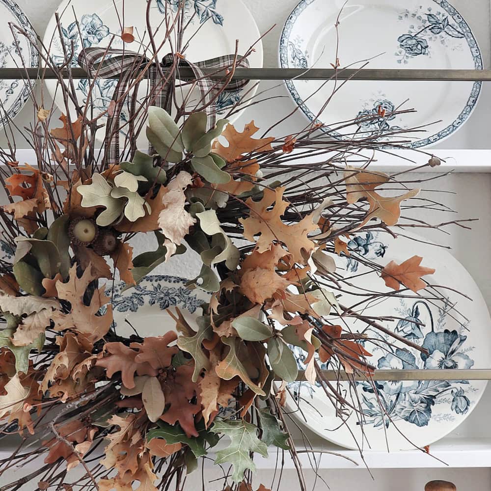A wreath made of dried leaves, twigs, and acorns hangs in front of a shelf displaying vintage white plates with blue floral patterns.