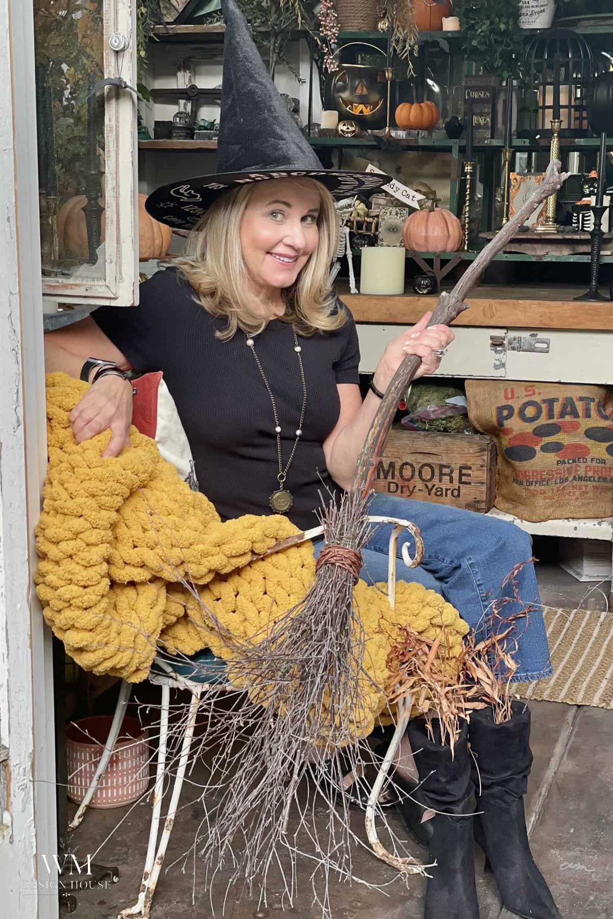 A smiling woman in a witch hat sits on a chair with a yellow blanket, holding a broomstick. Autumn decorations, pumpkins, and rustic items are in the background.