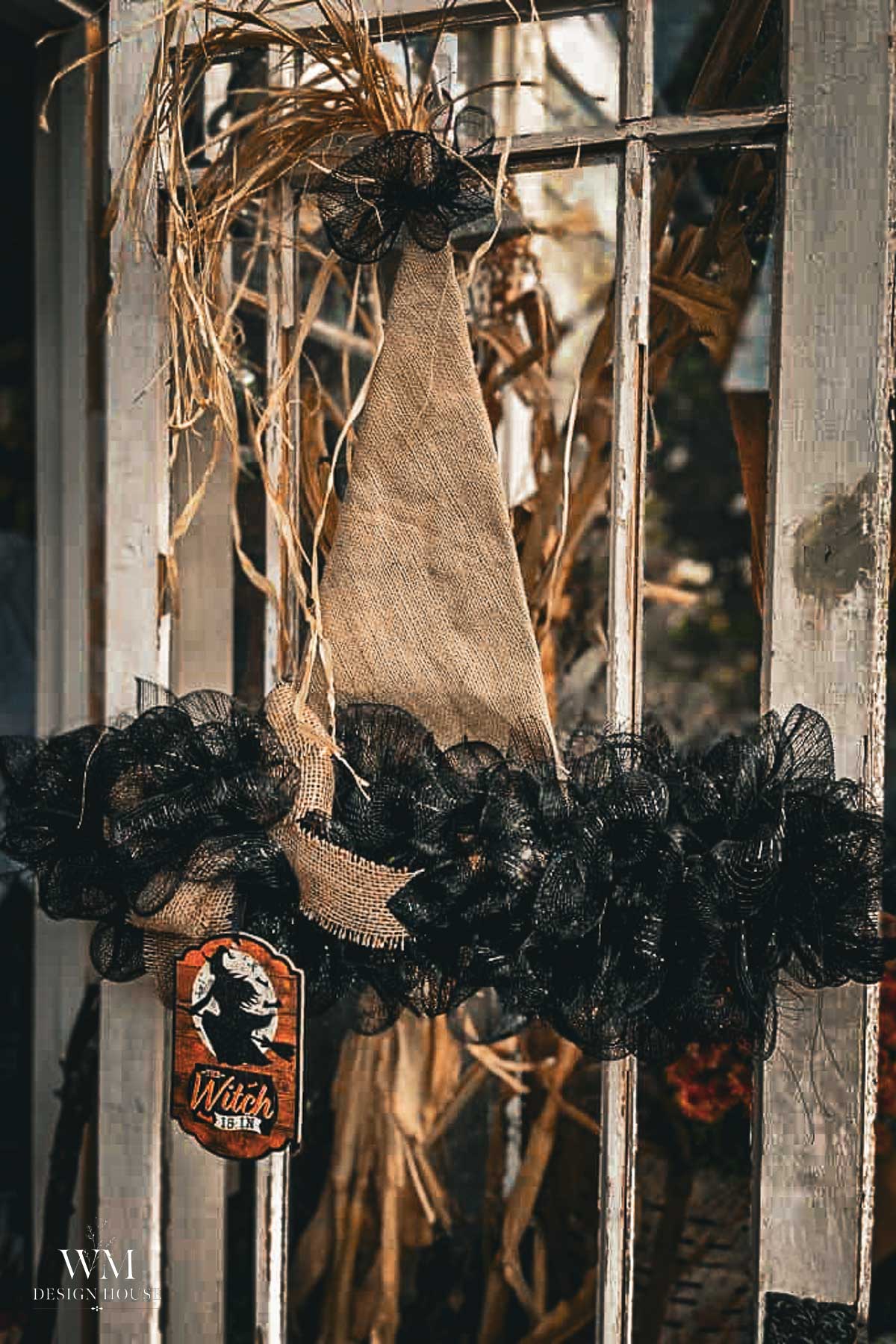 A rustic witch hat wreath with black mesh ruffles and a “Witch Inn” sign hangs on a glass door, with straw accents and corn stalks in the background.