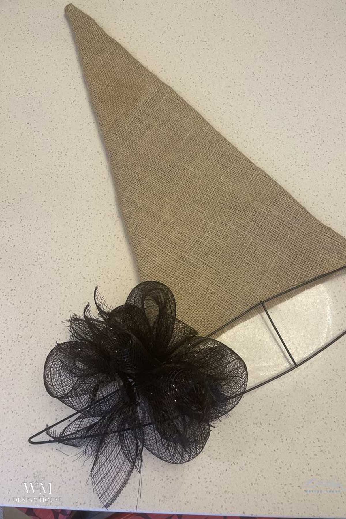 A handmade witch hat made of burlap with a wire brim and a large black mesh flower, placed on a speckled white surface.