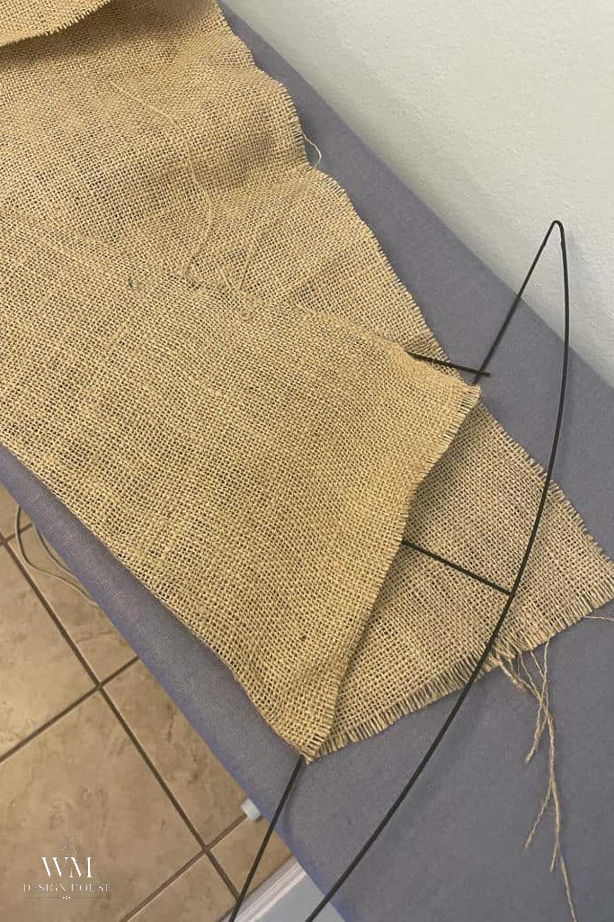 A piece of burlap fabric is draped over a black metal wreath frame on a gray surface, with tan tiles visible on the floor below.