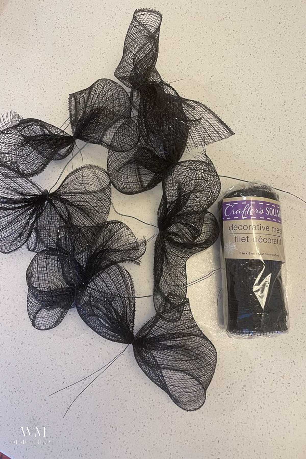 Black decorative mesh is cut and shaped into bow-like forms on a white surface. A roll of unopened black mesh labeled “Crafter’s Square decorative mesh” sits to the right of the finished shapes.