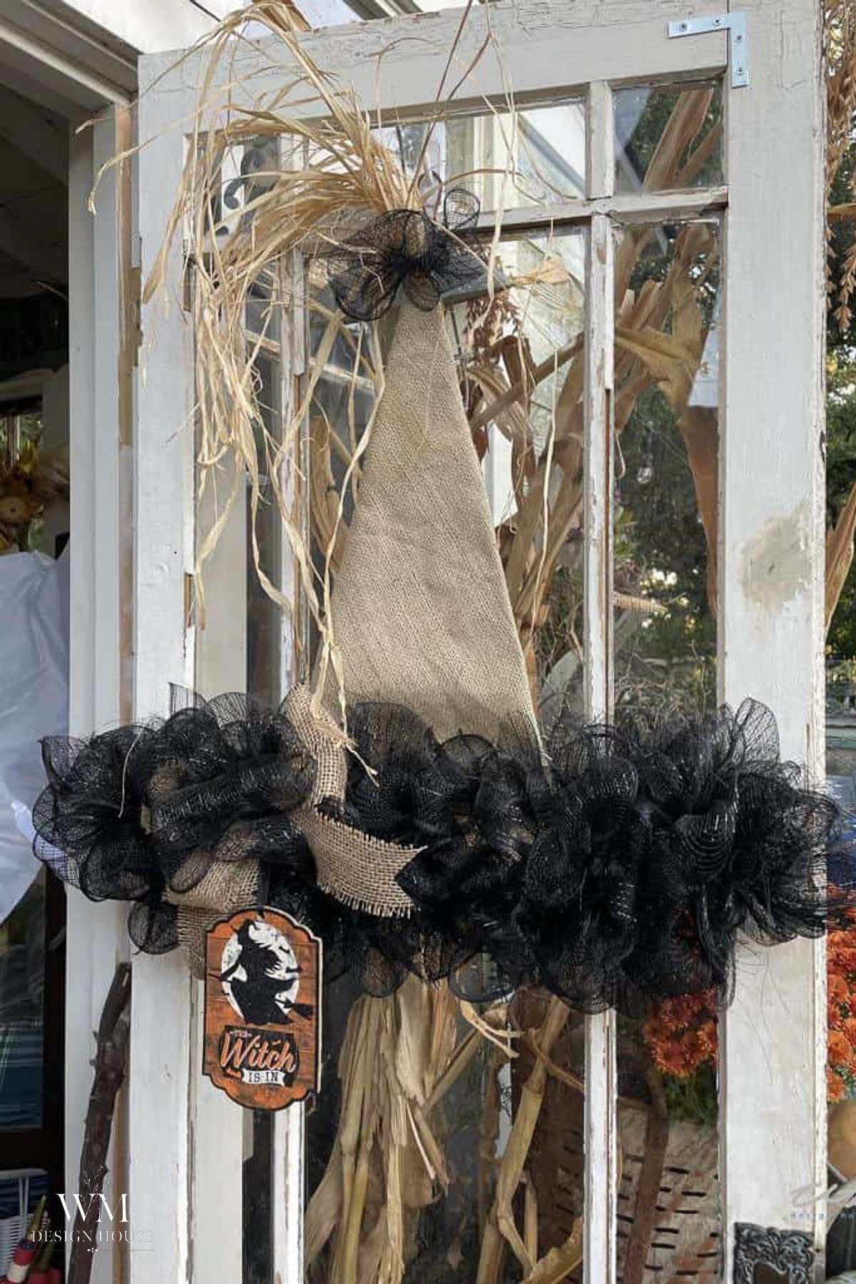 A witch hat wreath with black mesh, burlap, and straw hangs on a glass door. A small sign with a witch silhouette and the word “Witch” is attached at the bottom. Cornstalks are visible in the background.