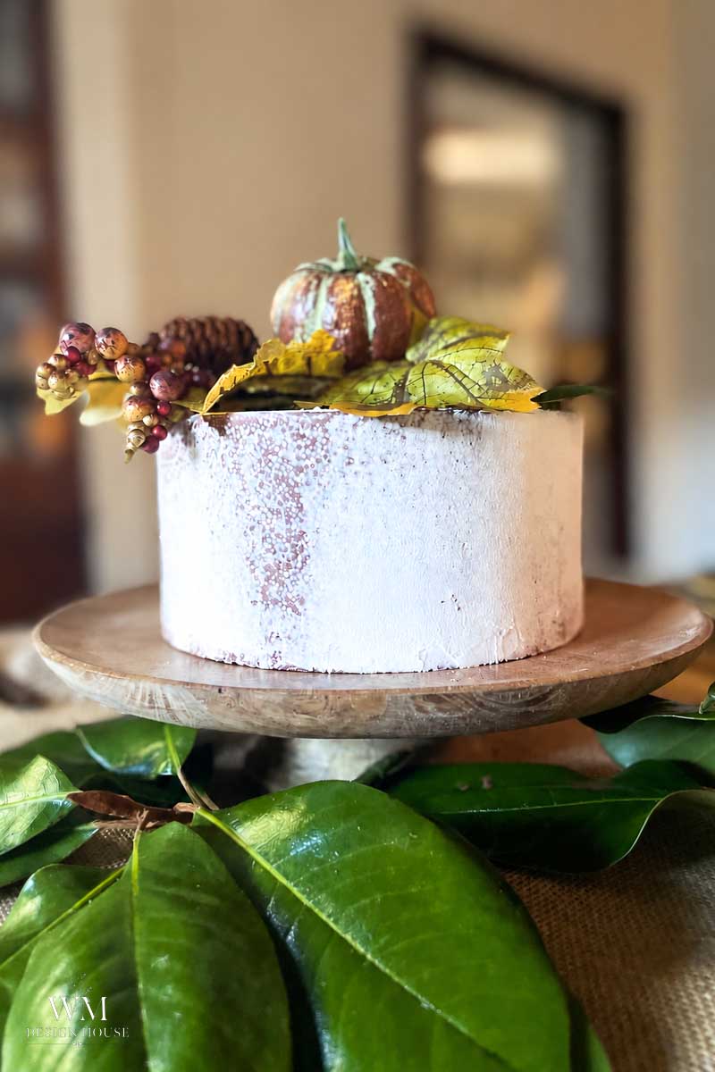 A rustic white frosted cake sits on a wooden stand, decorated with a small pumpkin, pinecones, berries, and autumn leaves, surrounded by green foliage—a charming centerpiece that embodies the spirit of an elegant fall tablescape.