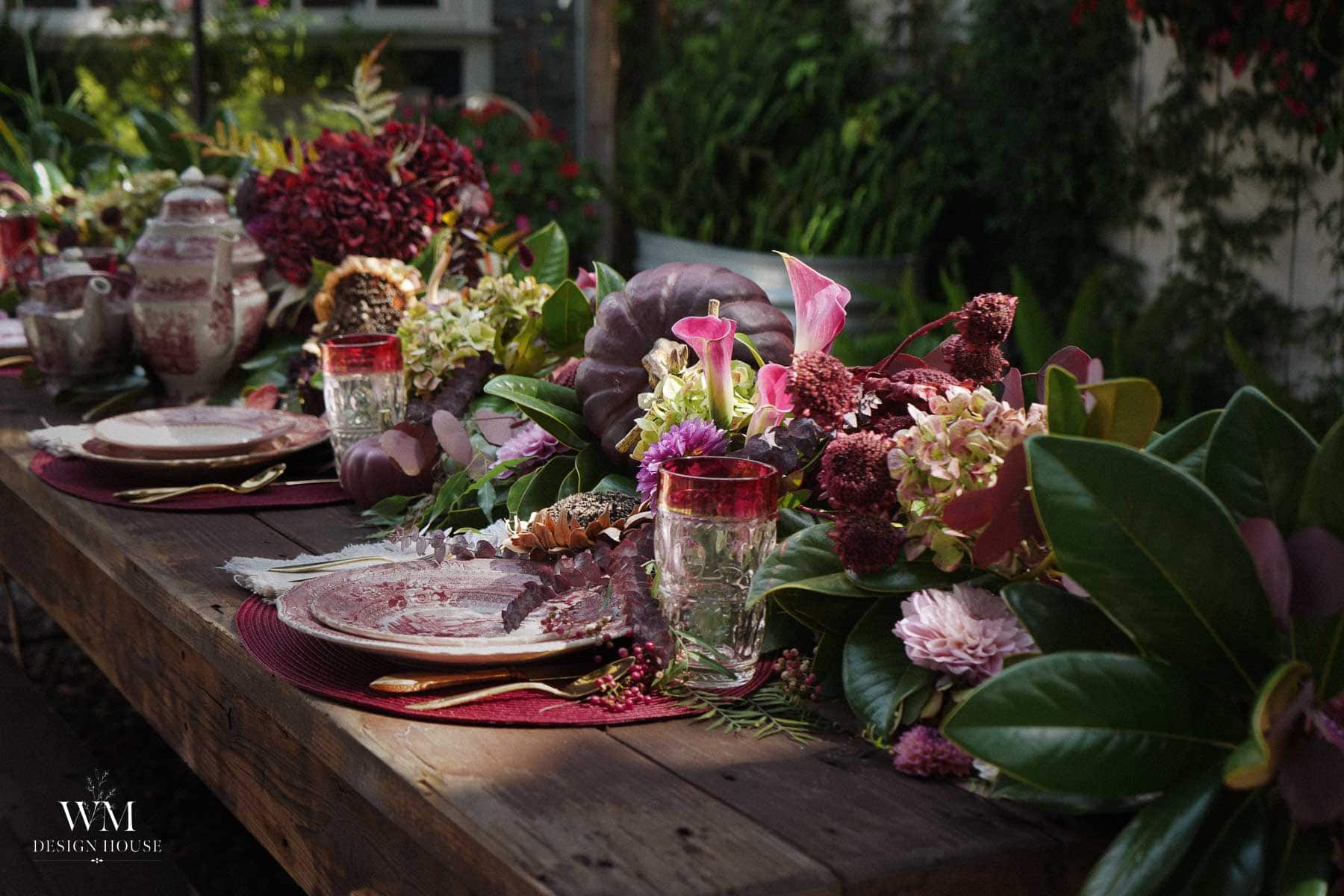 A rustic wooden table set for a meal with pink and red floral arrangements, vintage dishes, glasses with red rims, and teapots. Greenery and flowers create a lush, elegant centerpiece outdoors.