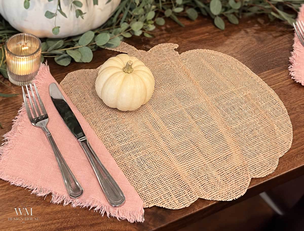 A pumpkin-shaped woven placemat with a small white pumpkin, pink napkin, fork, knife, and lit candle on a wooden table decorated with greenery creates an elegant tablescape perfect for showcasing your favorite fall tablescape ideas.