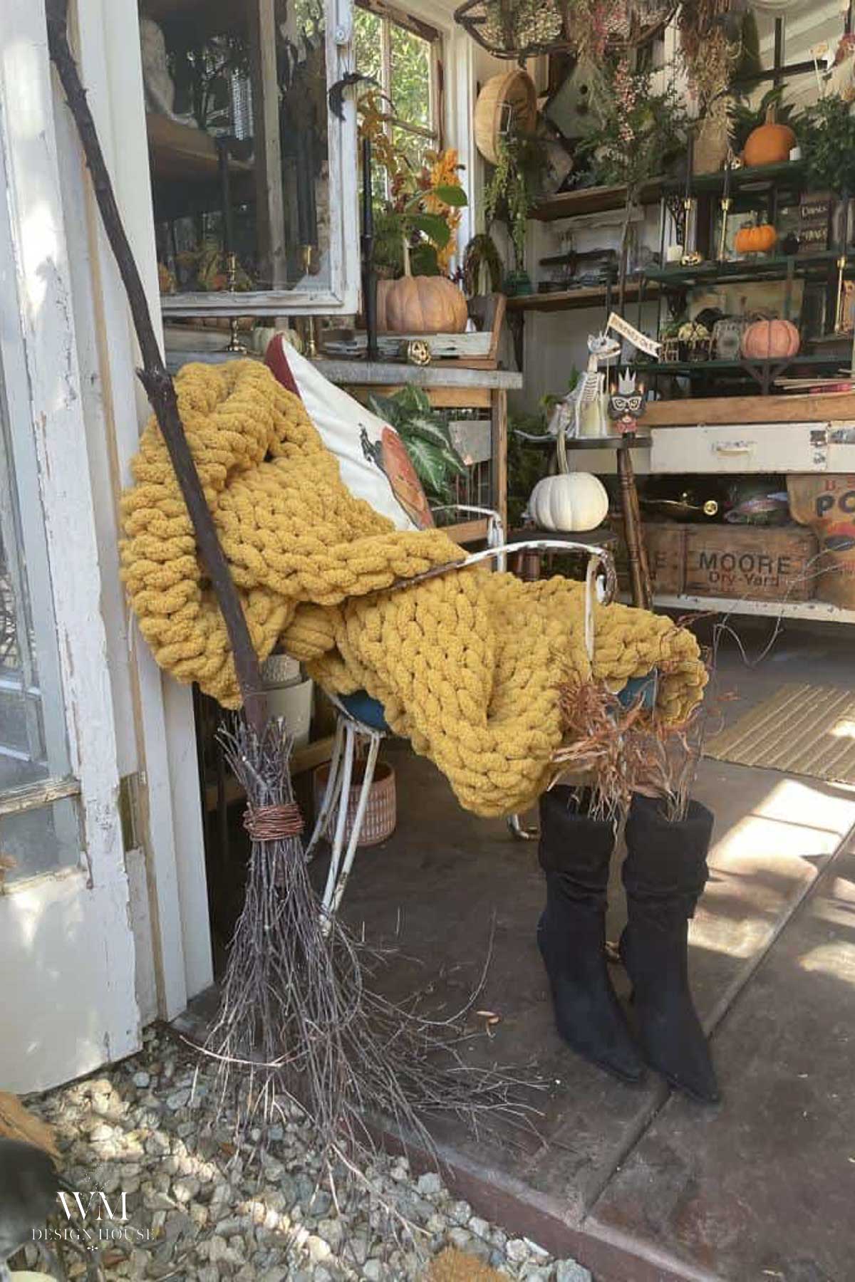 A cozy chair with a chunky yellow blanket, witch hat pillow, and black boots holding a broom sits in a rustic room decorated with pumpkins, plants, and gardening supplies.
