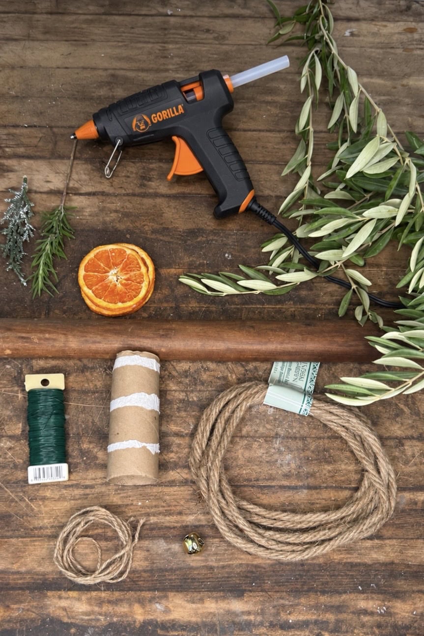 A glue gun, dried orange slice, olive branches, green wire, cardboard tube, rope, and a jingle bell laid out on a wooden surface to make Christmas napkin rings.
