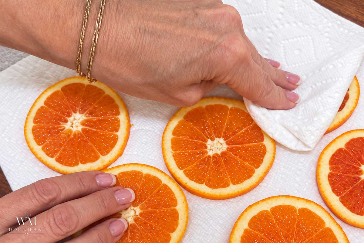 Hands blotting moisture from fresh orange slices with a paper towel as part of the process to make dehydrated orange slices.