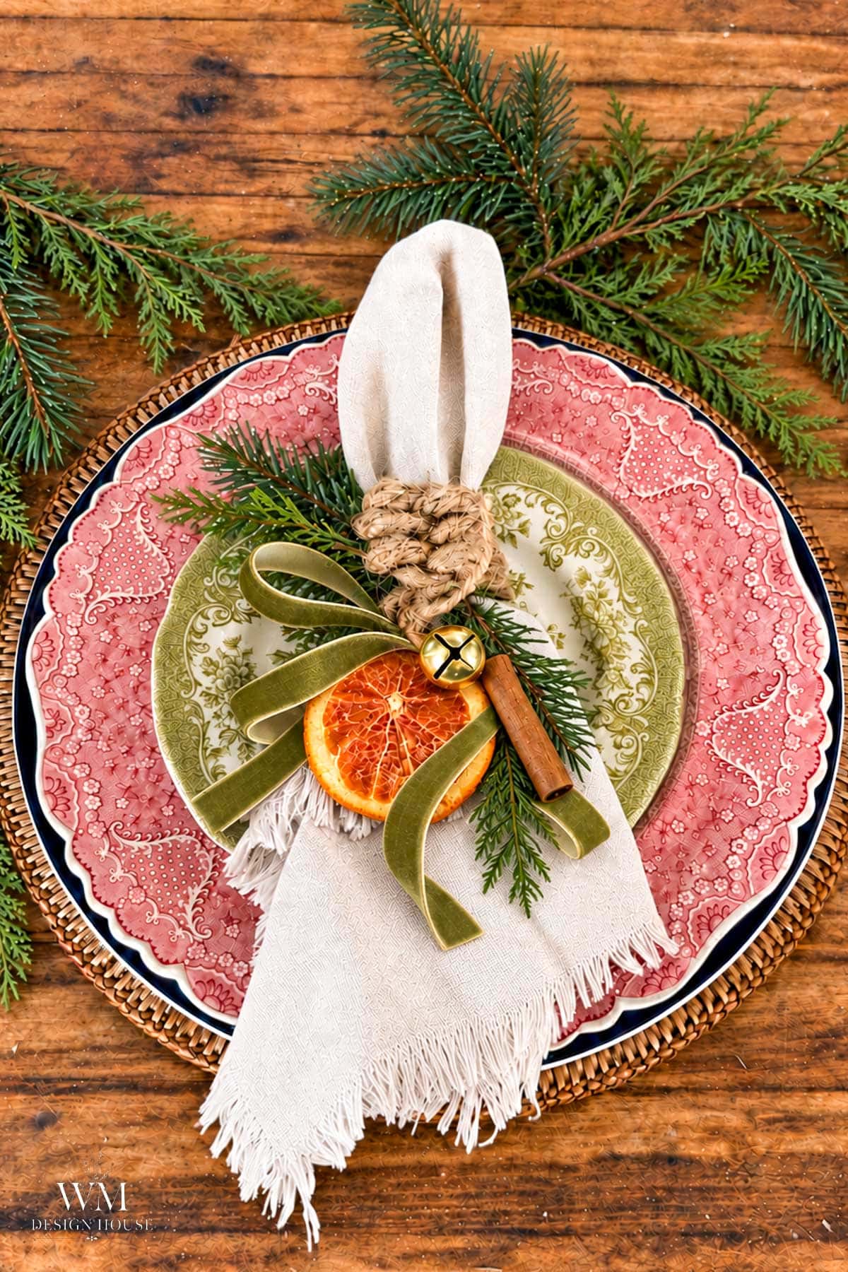 A place setting featuring a layered red and green patterned plate topped with a linen napkin, dried orange slices, a cinnamon stick, a small gold bell, and a green velvet ribbon tied with leaf knotted rope.