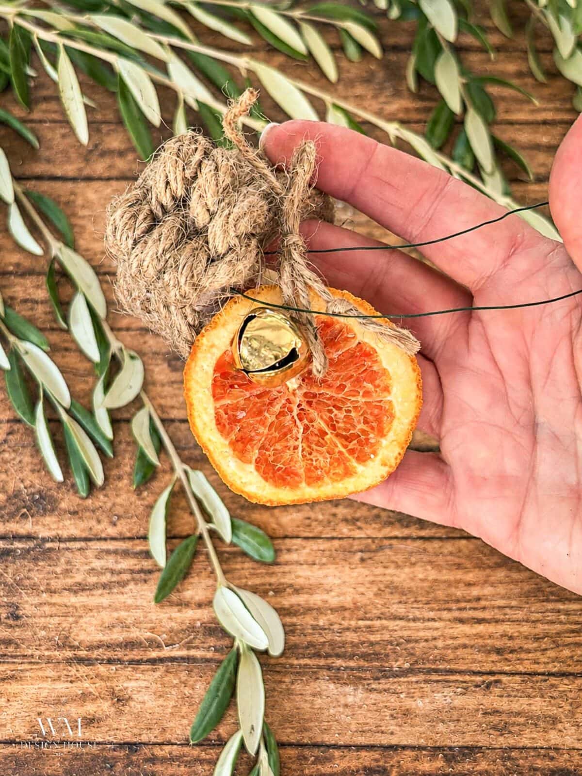 A hand holds a dried orange slice ornament decorated with a jute leaf knot and a small gold bell, resting against a wooden surface with scattered olive branches.