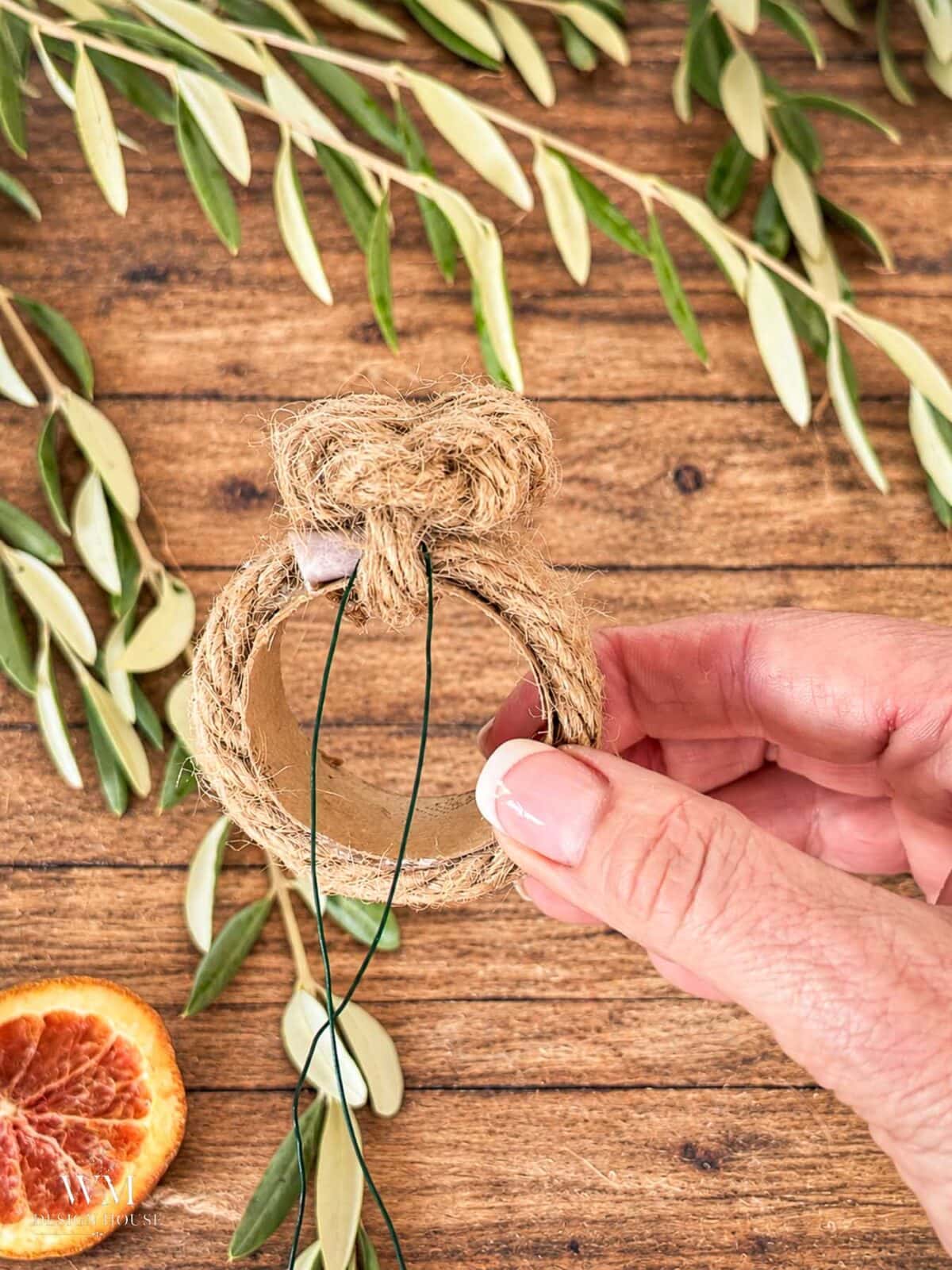 A hand holding a circular napkin ring made of wrapped jute rope and attaching a wire to add a dried orange slice. 