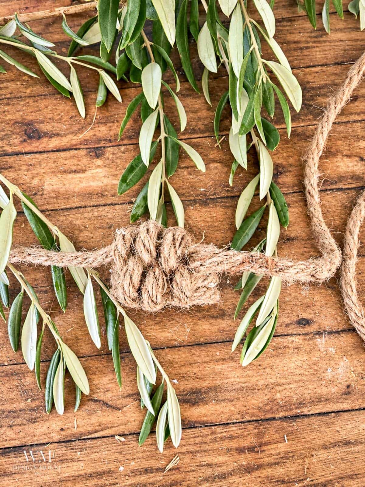 A  leaf knot of thick jute twine tied around a sprig of olive leaves on a wooden surface.