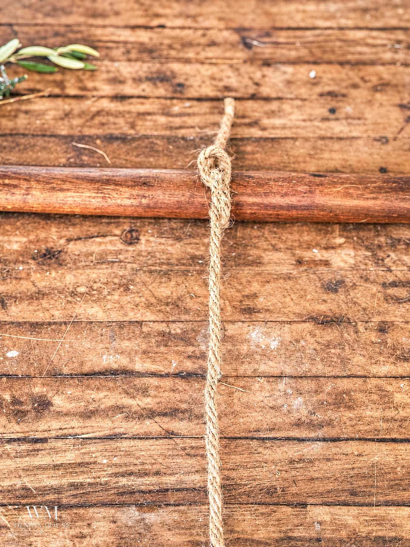 A length of twine tied in a knot around a horizontal broom handle against a rustic wood plank background.