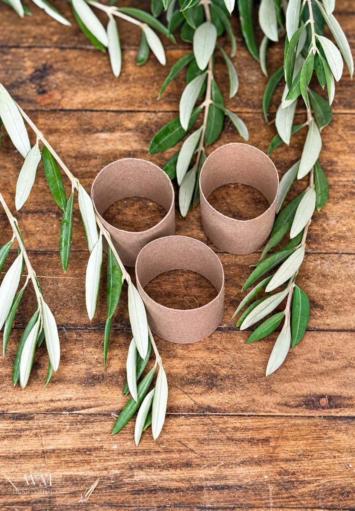 Three cardboard toilet paper rolls, repurposed as rustic Christmas napkin rings, are arranged on a wooden surface with olive branches.