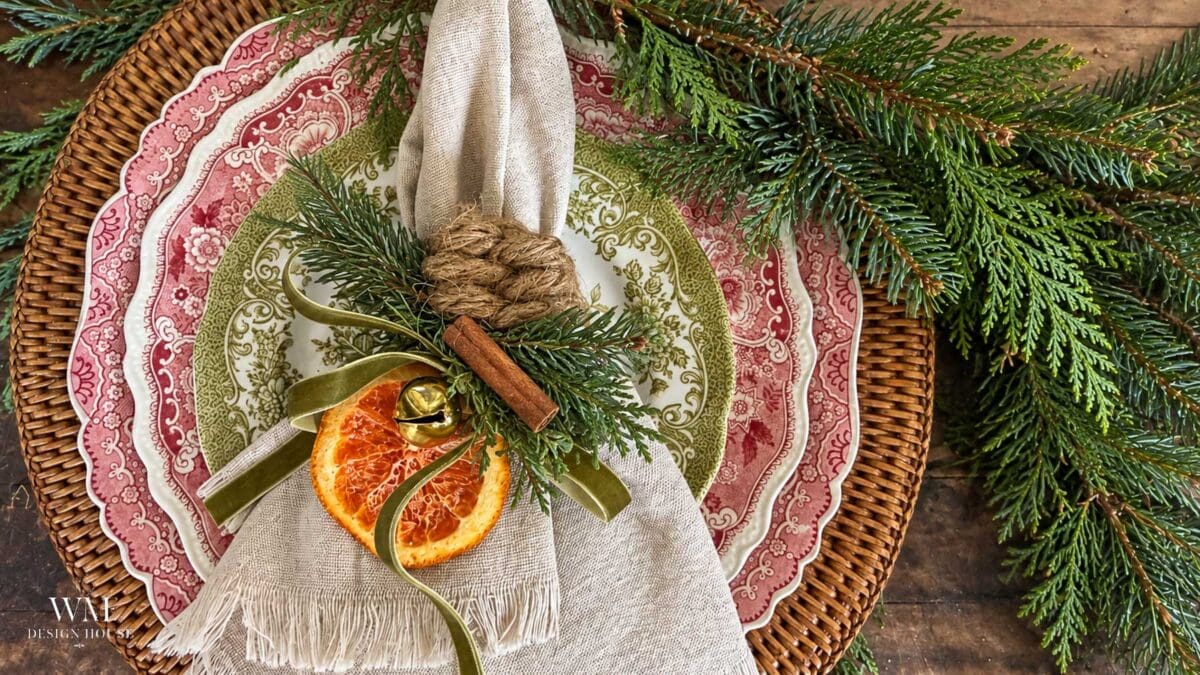 DIY Christmas napkin rings made with knotted jute rope, a dried orange slice, cinnamon stick, and evergreen on a red and green place setting.