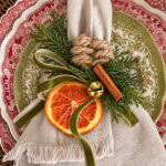 A beige linen napkin tied with a green velvet ribbon and decorated with a dried orange slice, a cinnamon stick, a small gold bell, and evergreen sprigs, resting on patterned plates.