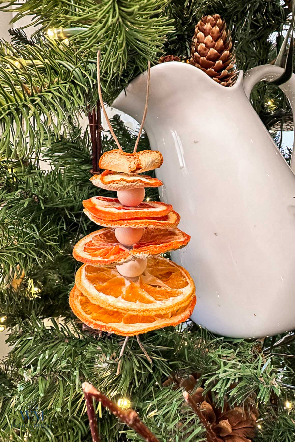 A handmade Christmas ornament made of stacked dried orange slices and wooden beads hanging on a pine tree next to a white ceramic pitcher.